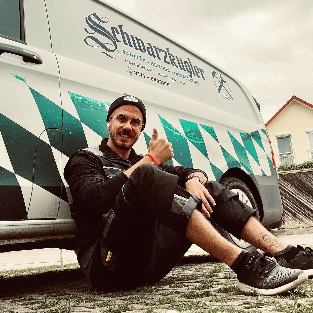 SHK Handwerker Michael Schwarzkugler sitzt auf dem Boden, angelehnt an ein Auto mit seiner Firmenaufschrift, zeigt Daumen hoch Geste.