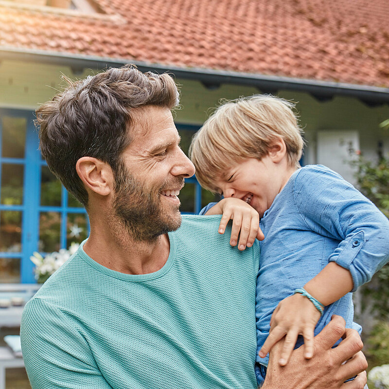 Vater mit lachendem Kleinkind vor Haus