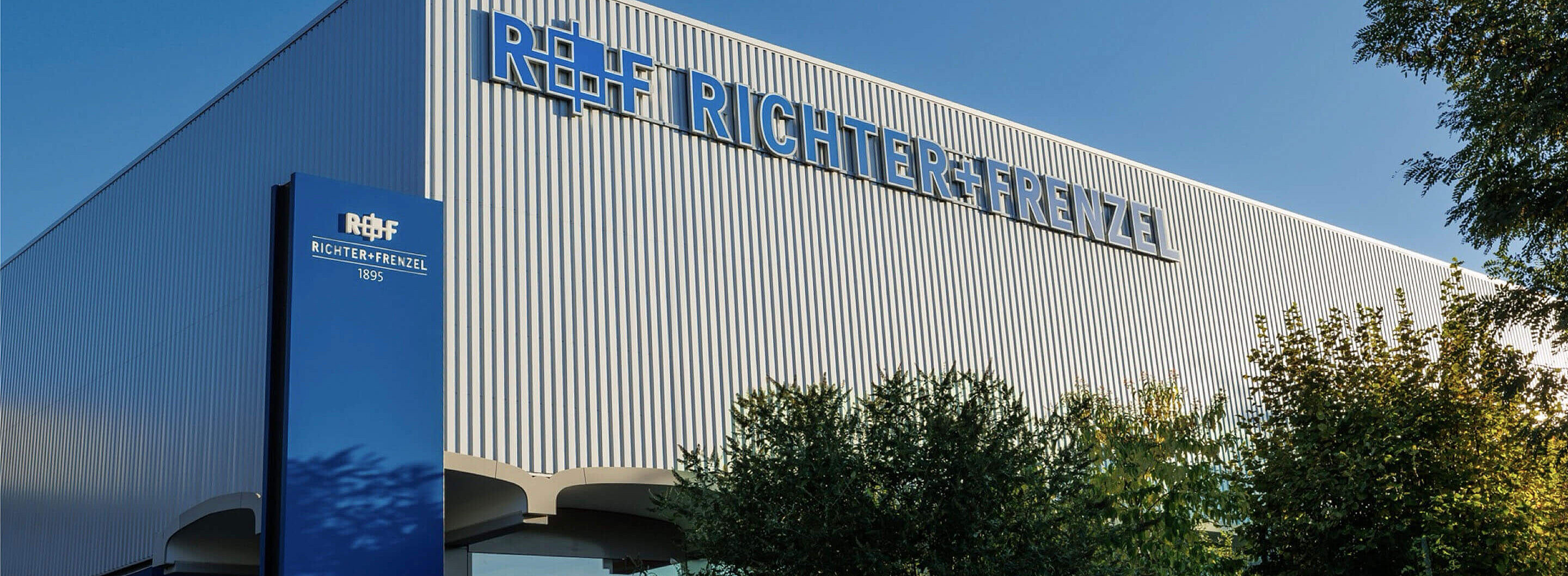 Fassade eines Unternehmensstandortes, darauf in blu  das R+F Logo sowie der Schriftzug Richter+Frenzel. Davor ein blauer Pylon mit der Aufschrift: R+F Richter+Frenzel 1895.