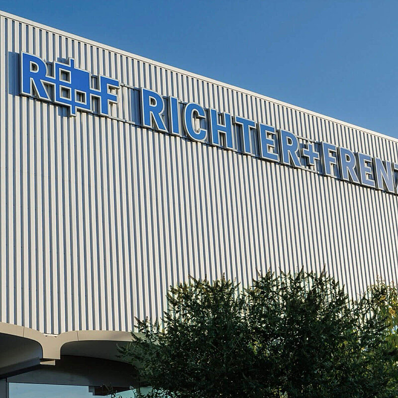 Fassade eines Unternehmensstandortes, darauf in blu  das R+F Logo sowie der Schriftzug Richter+Frenzel. Davor ein blauer Pylon mit der Aufschrift: R+F Richter+Frenzel 1895.
