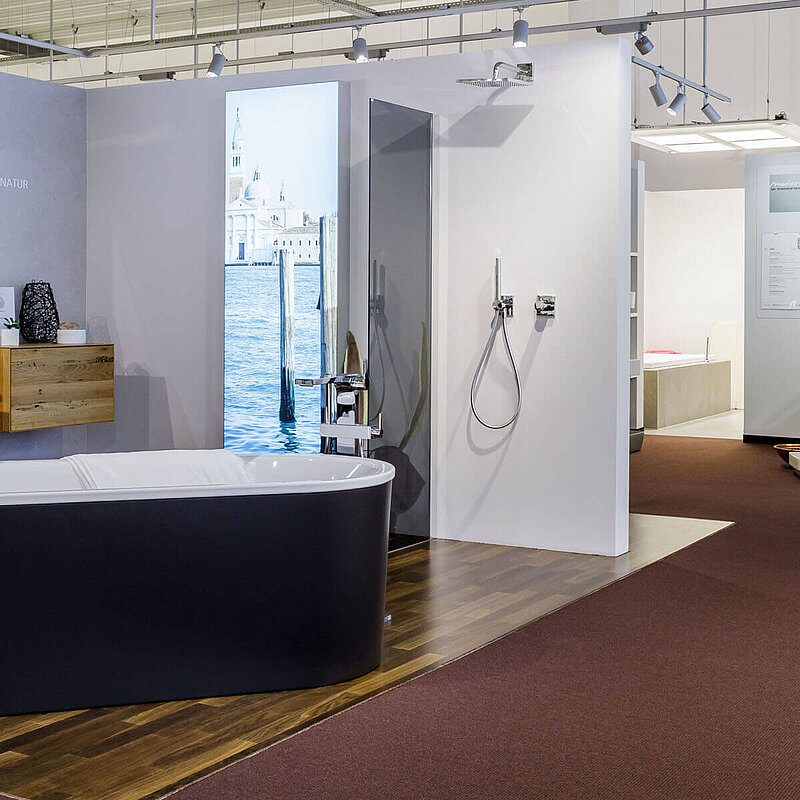 Blick in die Badausstellung in einem BäderStore. Im Vordergrund eine Badkoje mit freistehender schwarzer Badewanne, Doppelwaschtisch, Badmöbeln mit Holzfront sowei ein Dusche. Im Hintergrund sieht man Ausschnitte aus weiteren Badkojen.