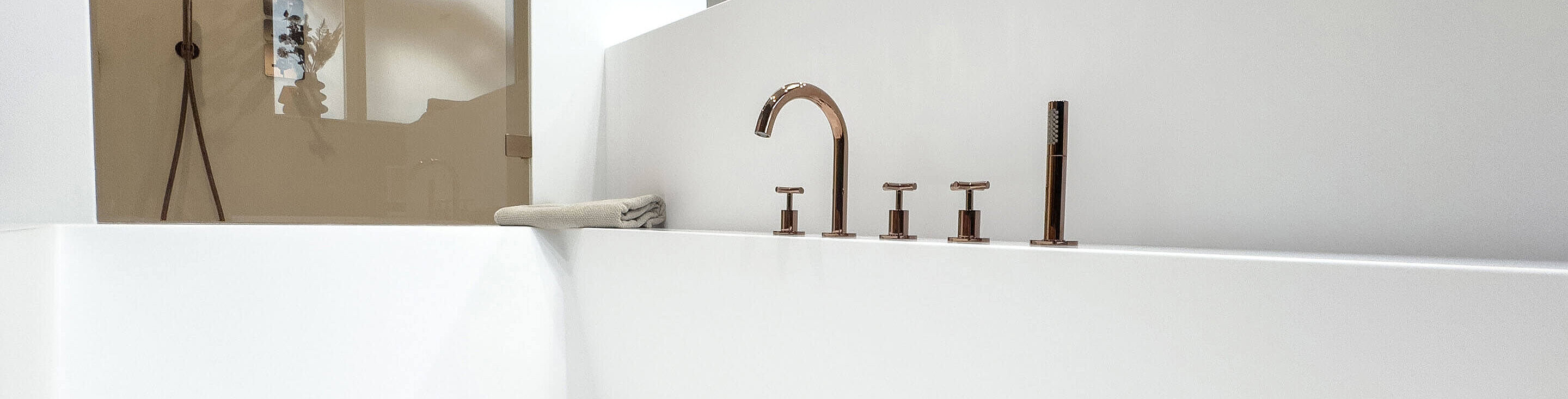 Nahansicht einer farbigen Badewannenarmatur. Von der Badewanne ist nur ein Teil des Randes zu sehen, darauf liegt ein Handtuch. Im Hintergrund eine Dusche mit Glastür und schwarzer Armatur.