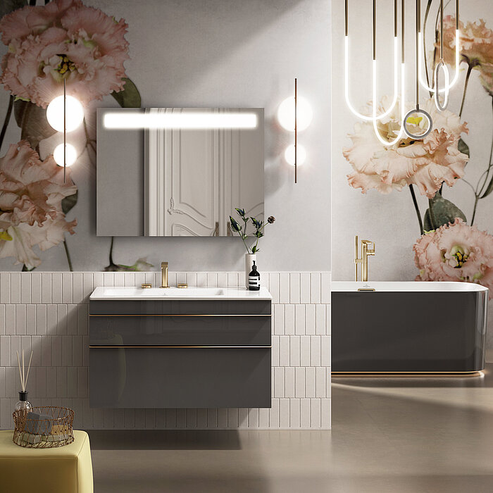 Ein modernes Badezimmer mit floral gemusterter Tapete in sanften Farben bildet den Hintergrund für einen grauen, wandhängenden Waschtisch mit goldenen Details und einem weißen Aufsatzwaschbecken samt goldener Armatur. Darüber hängt ein rechteckiger Spiegelschrank mit horizontaler Beleuchtung. Rechts im Bild ist eine elegante, ovale Badewanne in Dunkelgrau mit einer weiteren goldenen Armatur platziert. Moderne Pendelleuchten mit geschwungenen Formen und einzelnen Glühbirnen hängen über dem Waschbecken und der Badewanne und tragen zur stilvollen Atmosphäre des Raumes bei.